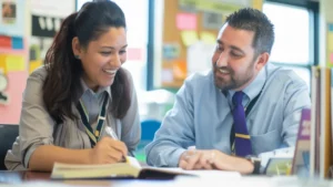 Two smiling teachers seated at a desk, looking at a textbook.